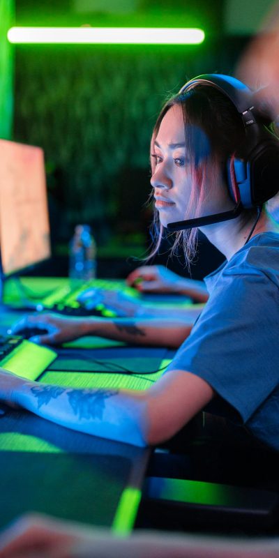 A woman deeply engaged in gaming, wearing headphones, with a visible tattoo and blue shirt.