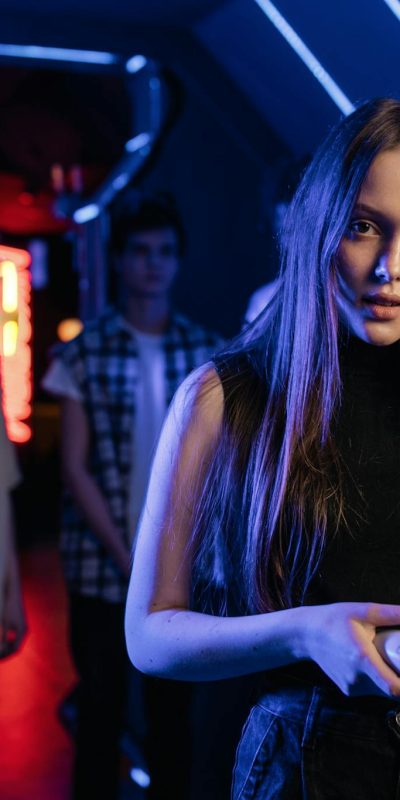 A young woman holding a game controller in a neon-lit arcade, surrounded by friends.
